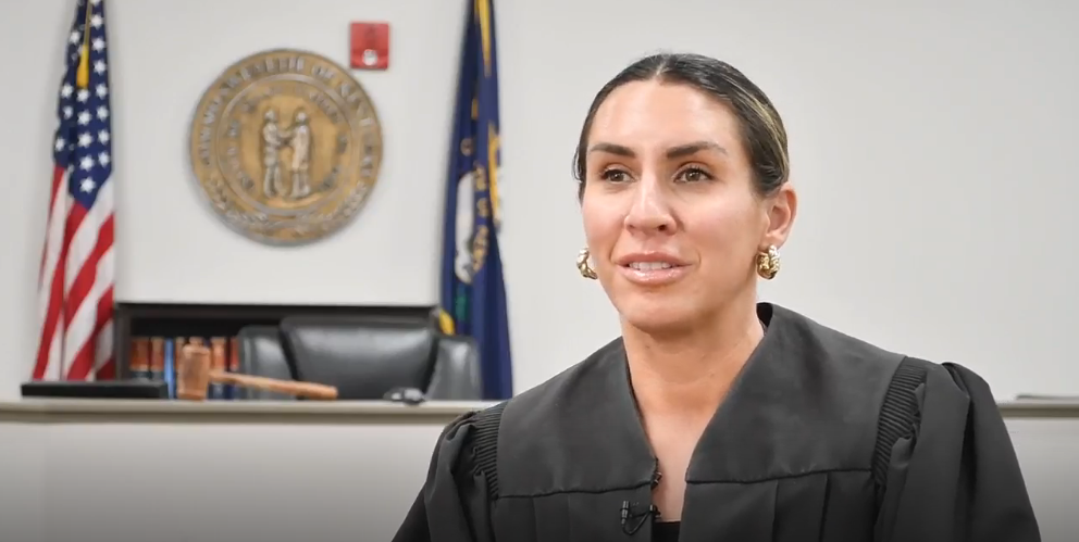 A female judge in a black robe speaks while seated in a courtroom, with the seal of the Commonwealth of Kentucky and state and U.S. flags visible in the background.